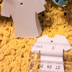 May include: A stack of white, blank, t-shirt shaped gift tags with a hole punched at the top. The tags are measured with a ruler, showing they are about 2.5 cm wide.