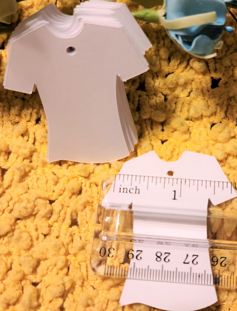 May include: A stack of white, blank, t-shirt shaped gift tags with a hole punched at the top. The tags are measured with a ruler, showing they are about 2.5 cm wide.