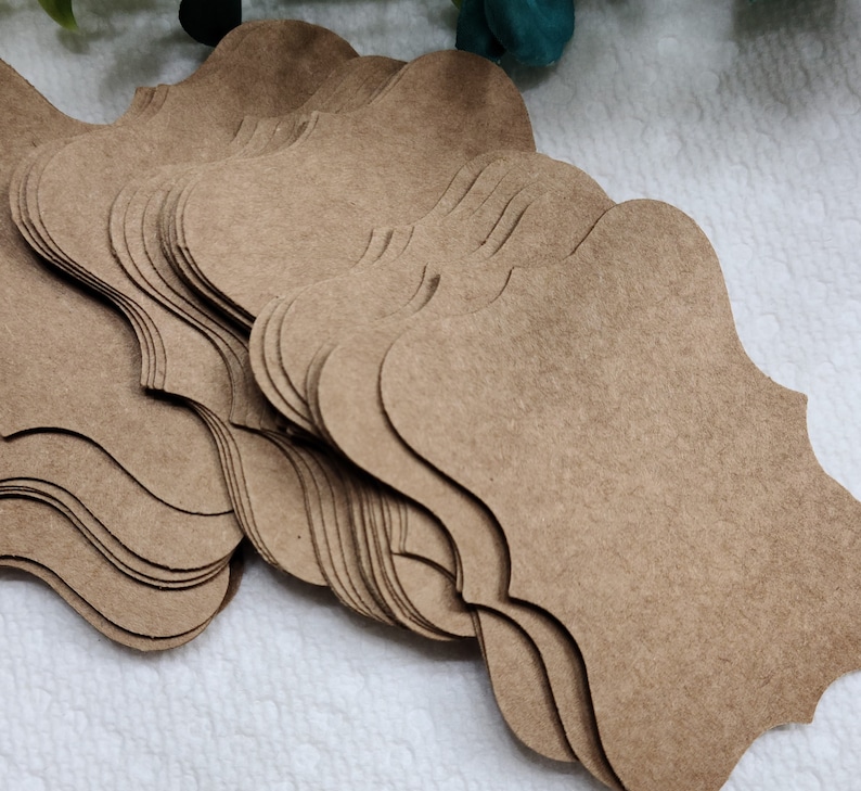 K&ouml;nnte beinhalten: Ein Stapel aus mehreren braunen Kraftpapier-Anh&auml;ngern, die jeweils eine dekorative Form mit gewellten R&auml;ndern und geschwungenen Seiten aufweisen. Die Anh&auml;nger bestehen aus flachem, mattem Karton und sind f&auml;cherf&ouml;rmig &uuml;bereinandergelegt.