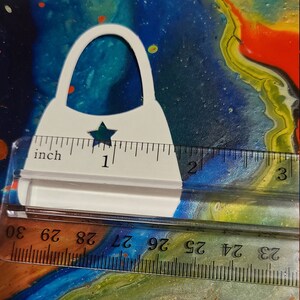 May include: White paper cutout of a handbag with a star in the center. The handbag is approximately 1 inch tall. The cutout is placed on a ruler with inch markings.