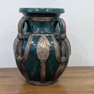 Może przedstawiać: Ciemnoturkusowy wazon ceramiczny z szerokim otworem i dwoma zakrzywionymi uchwytami. Zewnętrzna część jest ozdobiona dekoracyjnymi, metalicznymi pasami oraz owalnymi akcentami, które biegną od szyjki aż do podstawy naczynia.