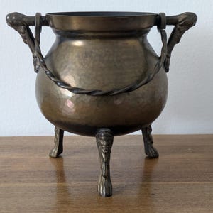 May include: A round, hammered brass cauldron with a dark patina finish. The vessel features three ornate legs with sculpted faces, two side handles with animal-head attachments, and a twisted metal swing handle that rests across the top rim.