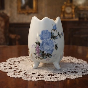 May include: A white ceramic vase with a scalloped rim, adorned with blue rose floral designs and three small feet. The vase is displayed on a white lace doily, set on a dark wooden table.