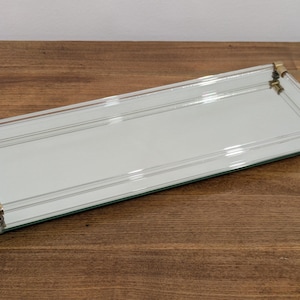 Op de afbeelding: Een rechthoekig spiegelplateau met een rand van helder glas en goudkleurige metalen accenten op de hoeken. Het plateau heeft een reflecterend oppervlak en een verhoogde glazen reling aan de lange zijden, ondersteund door goudkleurige hardware.