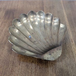 May include: A brass seashell-shaped dish with a scalloped design. The dish has a tarnished, aged appearance, with visible wear and discoloration. The dish is resting on a wooden surface.