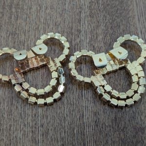 May include: Two gold-toned metal shoe clips featuring a circular design adorned with small, square-cut clear rhinestones. Each clip has a hinged metal mechanism on the back for attachment, showcasing a reflective, metallic finish throughout.