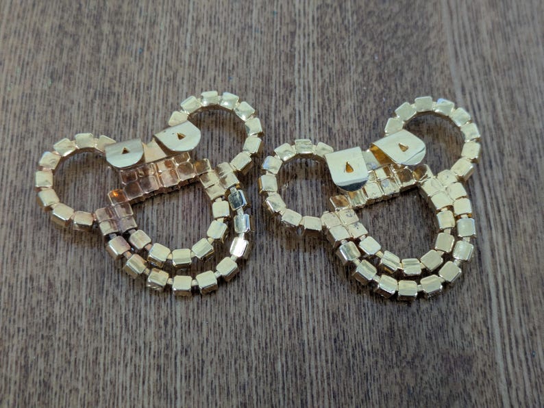 May include: Two gold-toned metal shoe clips featuring a circular design adorned with small, square-cut clear rhinestones. Each clip has a hinged metal mechanism on the back for attachment, showcasing a reflective, metallic finish throughout.