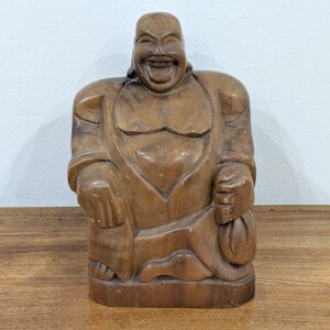 May include: A hand-carved wooden figurine of a laughing Buddha with a wide, open-mouthed smile, prominent ears, and a large belly. The figure is depicted in a seated position, wearing loose robes, and holding a small sack in the left hand.