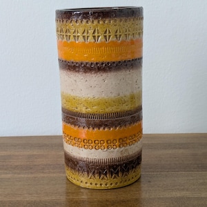 May include: A cylindrical ceramic vase featuring horizontal bands of brown, orange, yellow, and cream. The surface displays a textured, repeating geometric pattern of small circles and diamond shapes embossed into the clay across the coloured stripes.