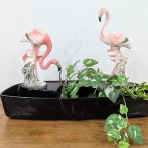 Puede incluir: Una jardinera de cerámica negra con diseño de varios niveles que presenta dos figuras de flamencos rosas y blancos. Un flamenco tiene la cabeza inclinada, mientras que el otro está erguido. Una planta verde y blanca variegada cuelga dentro.