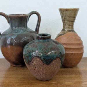 May include: Three ceramic vessels of varying shapes and sizes. One is a two-handled jug with a brown and teal glaze. Another is a small, round vase with a textured brown base and a glossy teal drip glaze. The third is a tall, ribbed vase.