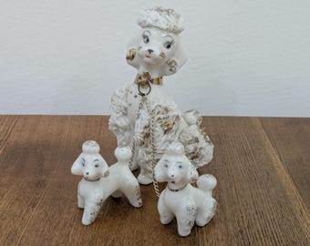 May include: A white ceramic poodle figurine set featuring one large poodle and two smaller poodles connected by thin silver-toned metal chains. Each poodle has gold-painted accents on its fur, ears, and tail, with painted facial features.