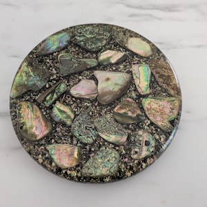 Puede incluir: Posavasos redondo con diseño de mosaico. Presenta piezas iridiscentes de concha de abulón incrustadas en una resina oscura y moteada. Las conchas muestran tonos de verde, azul y rosa, creando un artículo decorativo único.