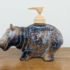 May include: A ceramic soap dispenser shaped like a hippopotamus with a glossy finish. The body features a mottled blend of deep brown, tan, and blue glaze. A light beige plastic pump mechanism is attached to the top of the hippo's back.