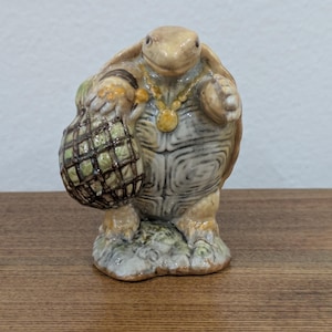 May include: A ceramic turtle figurine with a glossy finish, featuring a tan and grey shell, a yellow beaded necklace, and a small, round, green and brown chequered sack held in one hand, standing on a textured, mottled base.