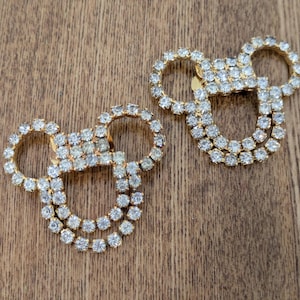 May include: Two gold-toned metal embellishments shaped like mouse heads, each featuring a double-row outline of clear, sparkling rhinestones. These decorative pieces have a circular head and two circular ears, creating a distinct silhouette.