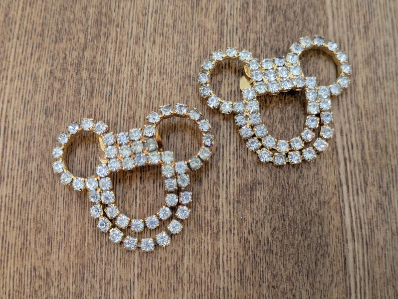 May include: Two gold-toned metal embellishments shaped like mouse heads, each featuring a double-row outline of clear, sparkling rhinestones. These decorative pieces have a circular head and two circular ears, creating a distinct silhouette.