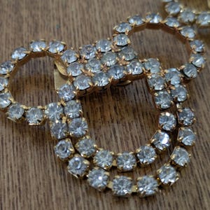 May include: A gold-toned metal brooch featuring a series of interconnected circular shapes. The entire surface of the metal frame is densely encrusted with small, clear, faceted rhinestones that reflect light, creating a sparkling appearance.