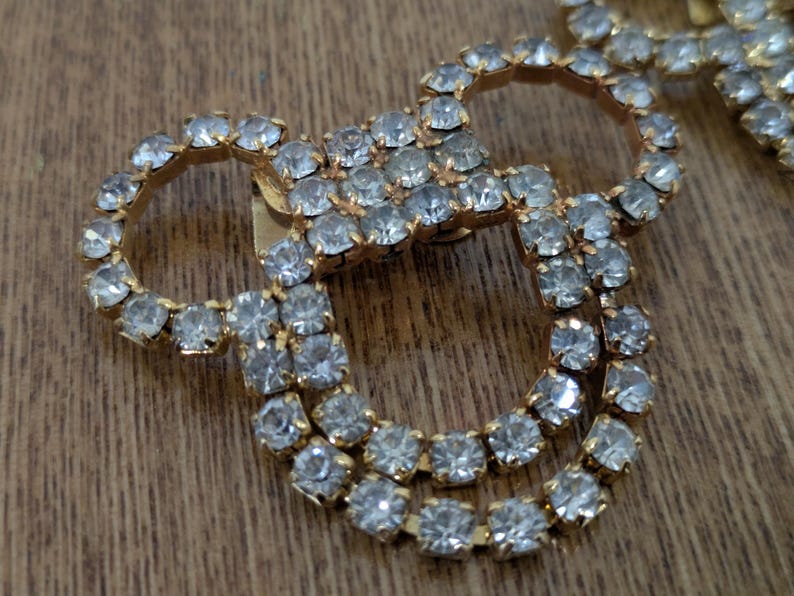 May include: A gold-toned metal brooch featuring a series of interconnected circular shapes. The entire surface of the metal frame is densely encrusted with small, clear, faceted rhinestones that reflect light, creating a sparkling appearance.