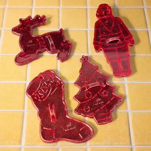 May include: Four red plastic Christmas cookie cutters in the shapes of a reindeer, Santa Claus, a Christmas stocking, and a Christmas tree. The cutters are on a yellow tiled surface.