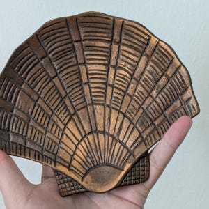 May include: A bronze-toned metal dish shaped like a scallop shell. The surface features a textured, radiating pattern of raised lines and grooves that mimic the natural ridges of a seashell, with a small grid-like pattern at the base hinge.