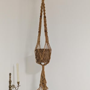May include: A tan macrame plant hanger featuring knotted cord construction, integrated wooden beads, and a woven basket container. The hanger terminates in a long, knotted tassel at the bottom and is suspended from a black metal ceiling hook.