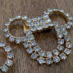 May include: A gold-toned metal brooch featuring a cluster of clear, faceted rhinestones arranged in a circular pattern. The design consists of a central square section connected to two larger circular loops, all encrusted with sparkling stones.