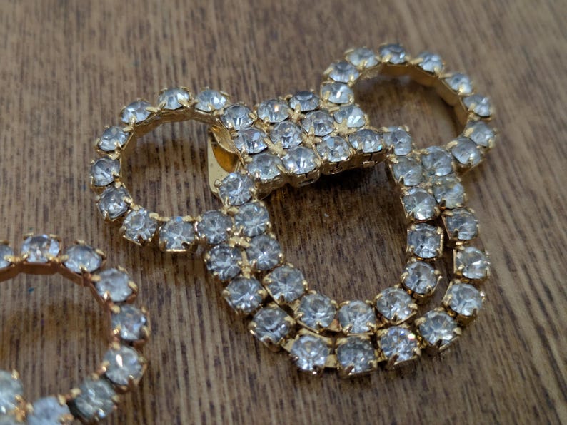 May include: A gold-toned metal brooch featuring a cluster of clear, faceted rhinestones arranged in a circular pattern. The design consists of a central square section connected to two larger circular loops, all encrusted with sparkling stones.