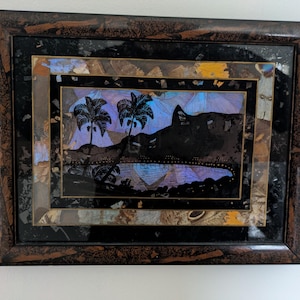 May include: Framed artwork featuring a tropical landscape silhouette with palm trees and a mountain against a blue and purple sky. The artwork is framed in a dark brown, textured frame with a multi-layered design.