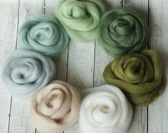 May include: A collection of seven different shades of green, beige, and white wool roving. The wool is arranged in a circle on a white wooden background.