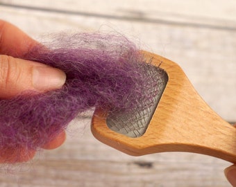 May include: A hand holds a strand of purple wool fibre over a wooden wool carder with metal teeth. The carder is used to align and blend fibres for spinning.
