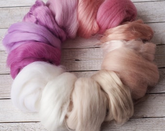 May include: A circle of colourful wool roving in shades of pink, purple, white, and beige. The roving is arranged in a gradient pattern, starting with the darkest shade of purple and ending with the lightest shade of beige.