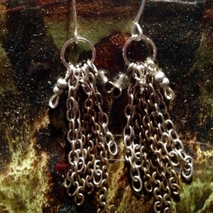 Puede incluir: Pendientes de cadena de plata con múltiples hebras de cadena que cuelgan de un aro de plata. Los pendientes están sobre un fondo marrón.