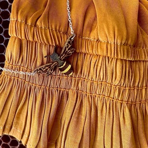 May include: A gold necklace with a wooden bee pendant hangs on a mustard yellow fabric with a gathered ruffle detail.