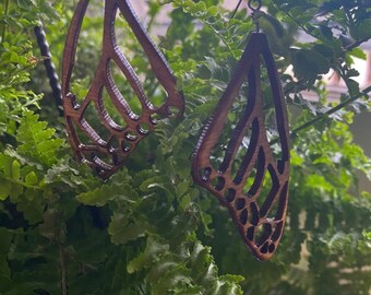 Puede incluir: Un par de pendientes de mariposa de madera con un acabado de madera marrón. Los pendientes están tallados intrincadamente con un diseño de mariposa detallado.