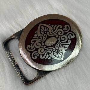 May include: A vintage metal belt buckle with an oval design. The buckle features a silver-colored filigree pattern on a dark red background. The buckle has a rectangular frame and a silver-colored finish.