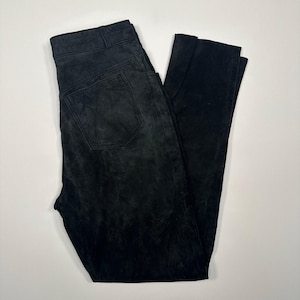 Può includere: Pantaloni neri in camoscio con un classico design a cinque tasche. I pantaloni hanno una chiusura con bottone e cerniera e passanti per cintura. Il tessuto ha un aspetto strutturato. I pantaloni sono piegati su una superficie bianca.