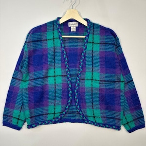 Könnte beinhalten: Ein Vintage-Cardigan mit offenem Schnitt in einem lebendigen Karomuster. Der Pullover hat eine Farbpalette aus Blau, Türkis und Lila mit einem geflochtenen Besatz an den Kanten. Das Kleidungsstück wird auf einem Holzbügel vor einem neutralen Hintergrund präsentiert.