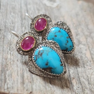 May include: A pair of silver earrings with a textured design. Each earring features a vibrant turquoise stone and a smaller, oval-shaped ruby. The earrings have a unique, handcrafted appearance.