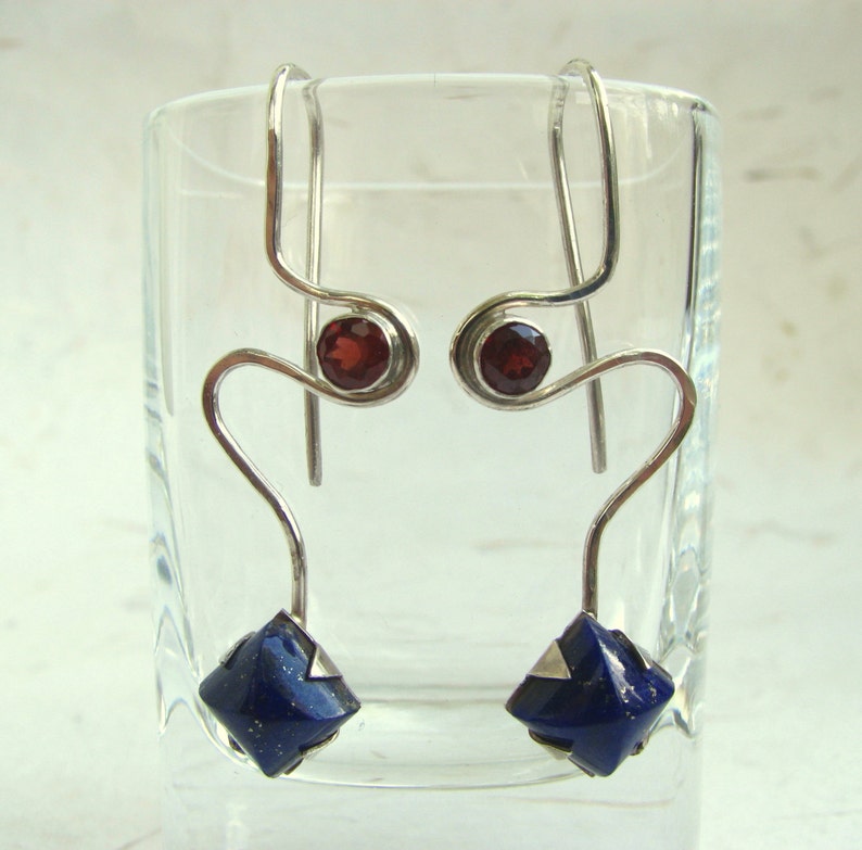 Puede incluir: Pendientes de plata con una piedra preciosa azul y una piedra preciosa roja. Los pendientes tienen forma de l&iacute;nea curva con un bucle en la parte superior.