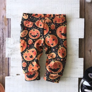 May include: Black baby pants with a repeating pattern of orange and yellow jack-o'-lanterns. The pants have a black background with orange and yellow pumpkins of various sizes. The cuffs are folded over.