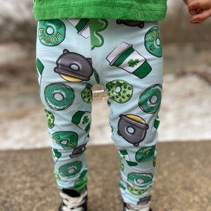 May include: Light blue leggings with a St. Patrick's Day theme. The leggings feature a repeating pattern of green donuts with the word "LUCKY", coffee cups with shamrocks, and pots of gold. The leggings are worn with a green shirt and black sneakers.