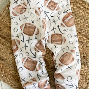 Puede incluir: Leggings blancos para bebé con un estampado repetido de balones de fútbol americanos marrones, flechas grises y equis y oes negras. Los leggings tienen cintura y puños elásticos.
