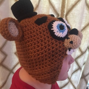 May include: A brown crocheted hat with a black top hat, a black eye patch, and a white and blue eye. The hat is shaped like a bear's head.