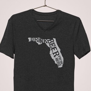 May include: A dark heather grey V-neck t-shirt with a white outline of the state of Florida. The text "Drink Beer From Here" is written vertically inside the outline of the state.