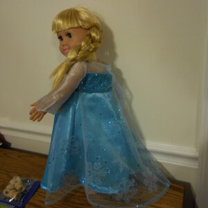 May include: A blonde doll wearing a blue and white dress with a sparkly design. The dress has a sheer overlay and a long train.