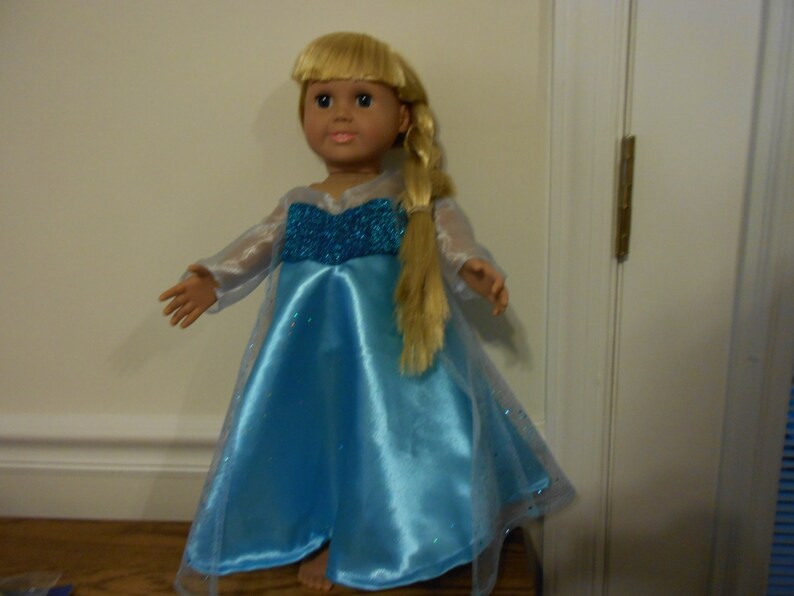 May include: A blonde doll wearing a blue and white dress with a sparkly bodice. The dress has a long, flowing skirt and a sheer overlay. The doll is standing on a wooden surface.