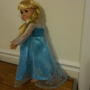 May include: A blonde doll wearing a blue and white dress with a snowflake design. The dress is made of a sheer fabric and has a long, flowing skirt.