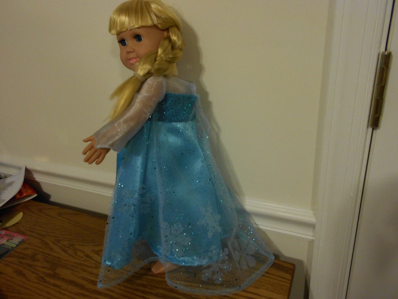 May include: A blonde doll wearing a blue and white dress with a snowflake design. The dress is made of a sheer fabric and has a long, flowing skirt.
