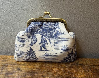 May include: A coin purse featuring a blue and white toile-style fabric print of a Bigfoot figure walking through a forest landscape. The purse has a brass-toned metal kiss-lock frame closure at the top and a structured, rounded rectangular shape.